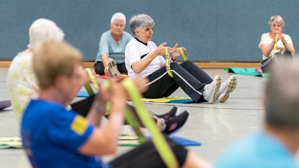 Senioren nehmen an einer Fitnessübung mit Fitnessbändern in einer Sporthalle teil, sitzend auf Fitnessmatten.