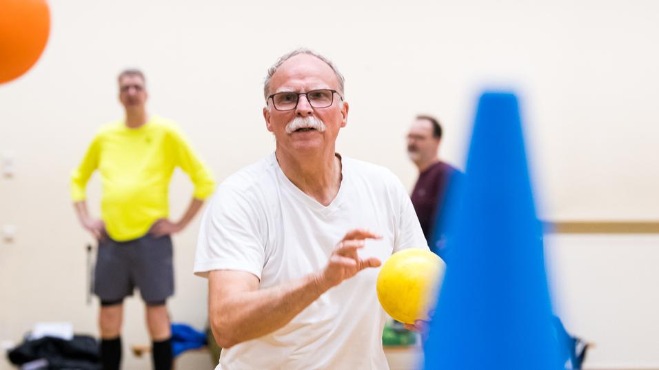 Mann mit Brille wirft einen gelben Ball in einem Sportraum, während andere Teilnehmer im Hintergrund stehen.
