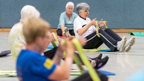 Senioren nehmen an einer Fitnessübung mit Fitnessbändern in einer Sporthalle teil, sitzend auf Fitnessmatten.