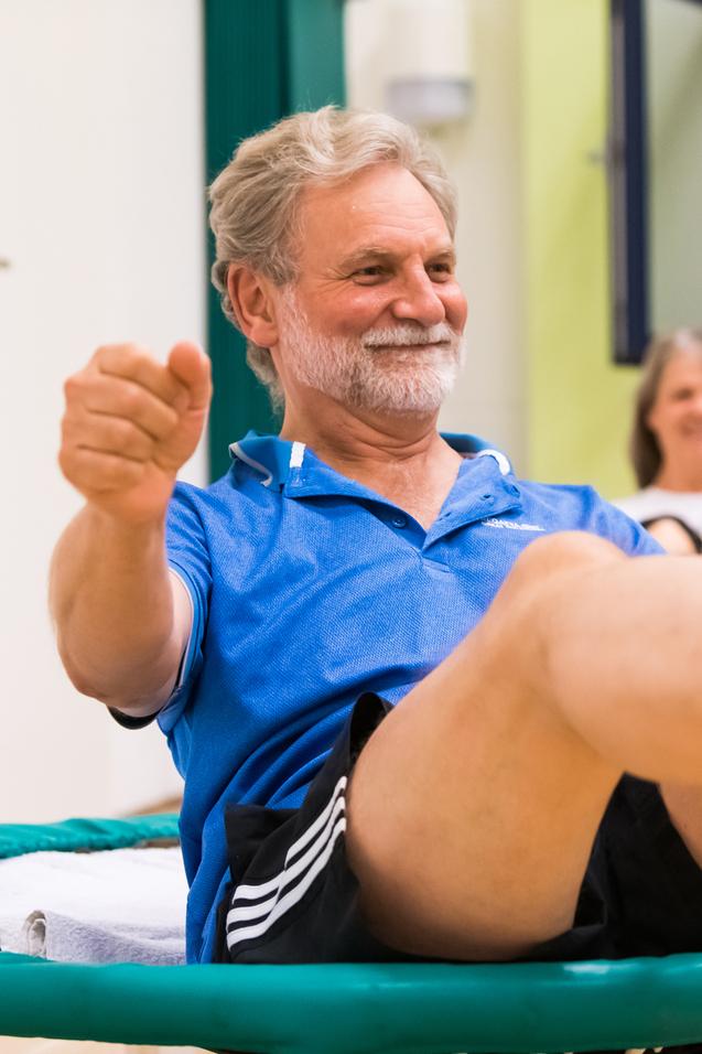 Älterer Mann macht Übungen auf einem Trampolin, lächelnd, mit einem anderen Teilnehmer im Hintergrund.