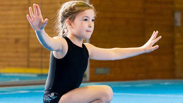 Zwei Mädchen in Gymnastikanzügen üben auf einer blauen Matte, eine kniet mit ausgestreckten Armen.